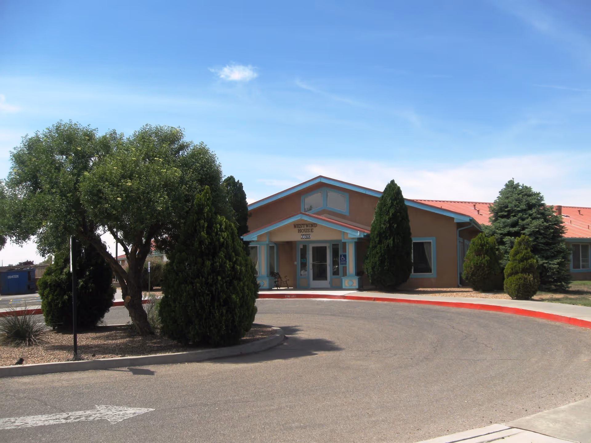 Front exterior view of Westwind House Assisted Living building with a curved driveway, several green trees and shrubs, and a clear blue sky.