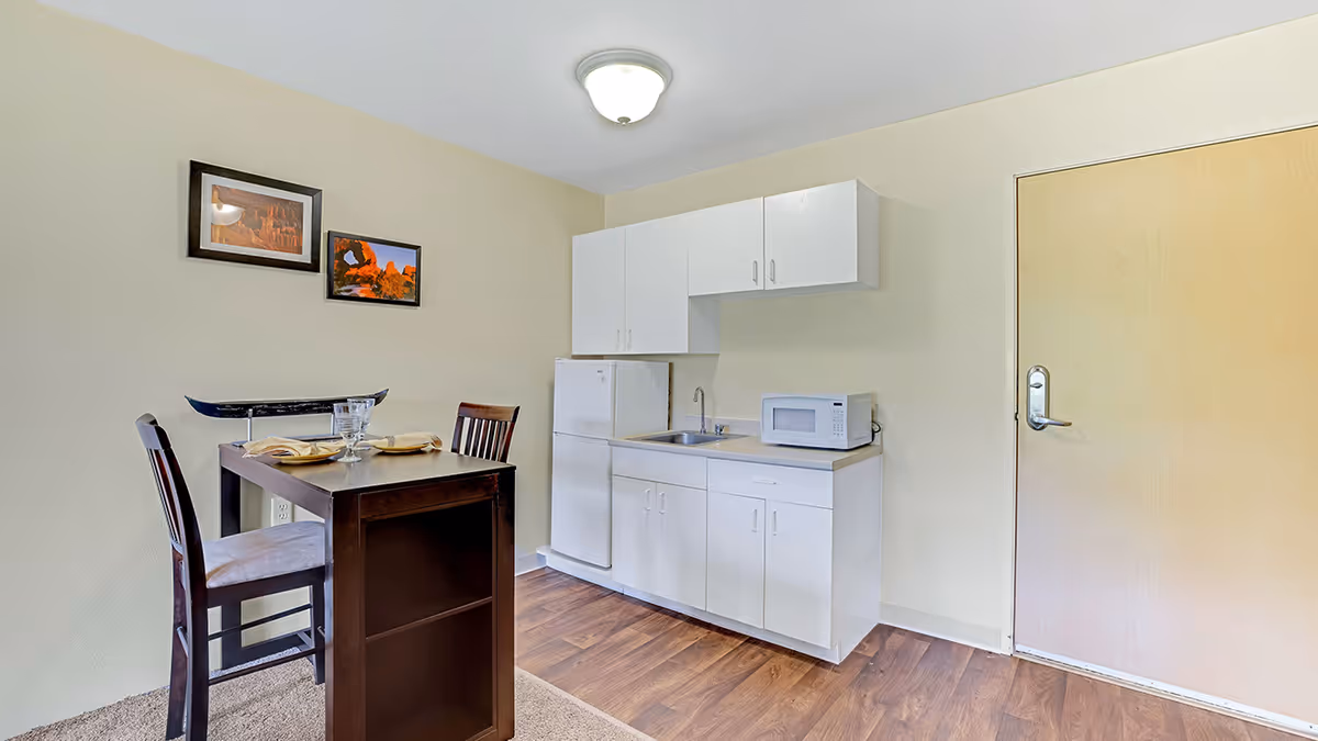 Small kitchenette area with white cabinets, a refrigerator, a microwave, and a sink. Next to the kitchenette is a small dining table set for two with two chairs. Two framed pictures hang on the beige wall above the table. The floor is a combination of carpet and wood, and there is a closed wooden door to the right.