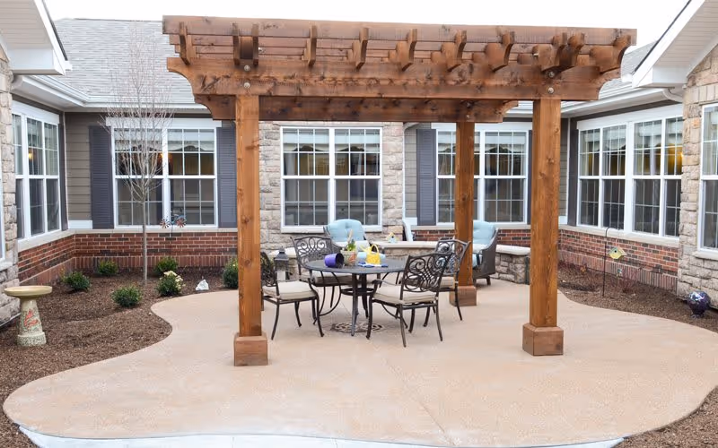 Outdoor patio area at Bickford of Aurora with a wooden pergola over a round table and four chairs. The patio is surrounded by a garden bed with small shrubs and a birdbath, and the building with multiple windows is visible in the background.
