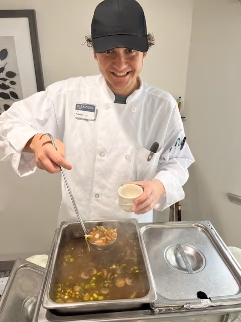 A smiling chef wearing a white coat and black cap ladles soup from a large metal container into a small bowl in a kitchen setting.