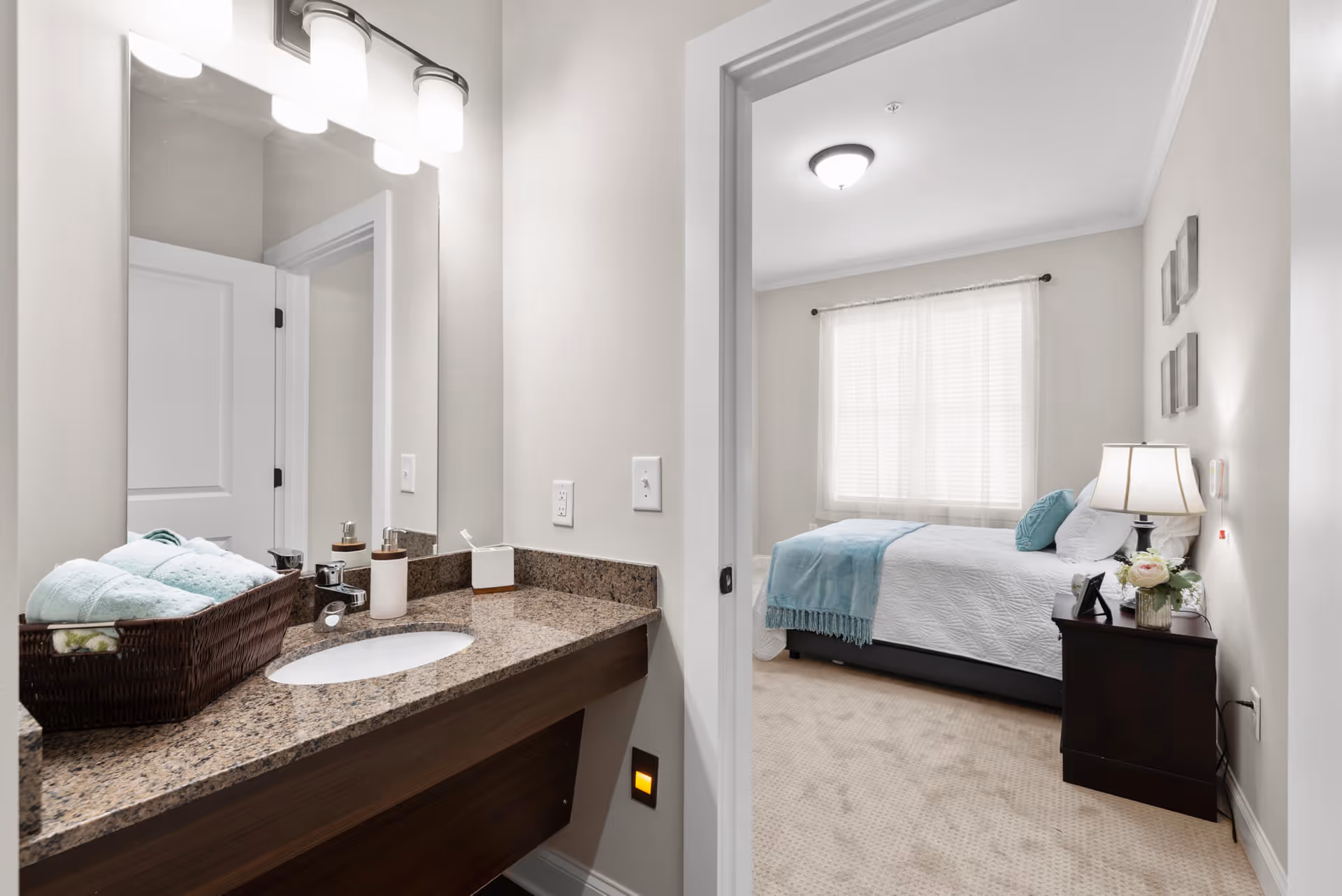 View of a bathroom vanity with a granite countertop, sink, and mirror on the left side, with a basket of folded towels and soap dispensers. Through an open doorway, a bedroom is visible with a neatly made bed featuring white bedding and a light blue throw, a nightstand with a lamp and flowers, and a window with sheer curtains.