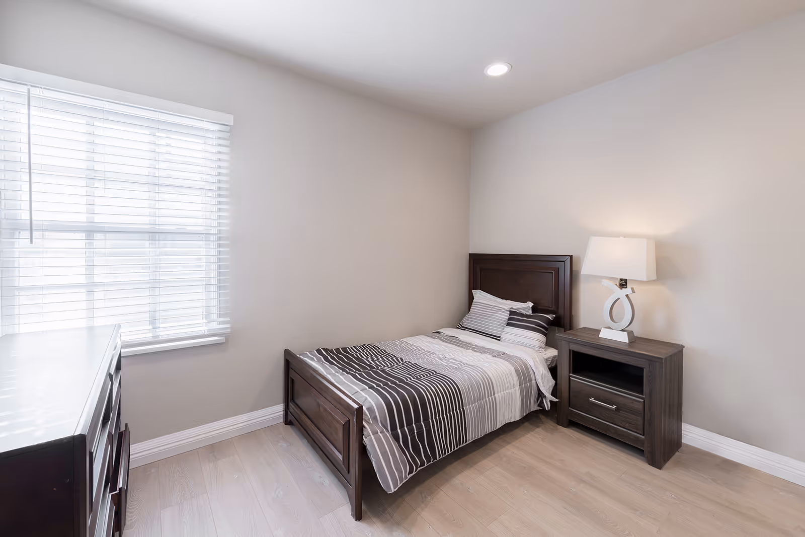A small bedroom with a single bed featuring striped bedding in shades of gray, white, and black. Next to the bed is a wooden nightstand with a modern white lamp. There is a window with white blinds on the left wall and a wooden dresser partially visible in the foreground. The room has light-colored walls and light wood flooring.