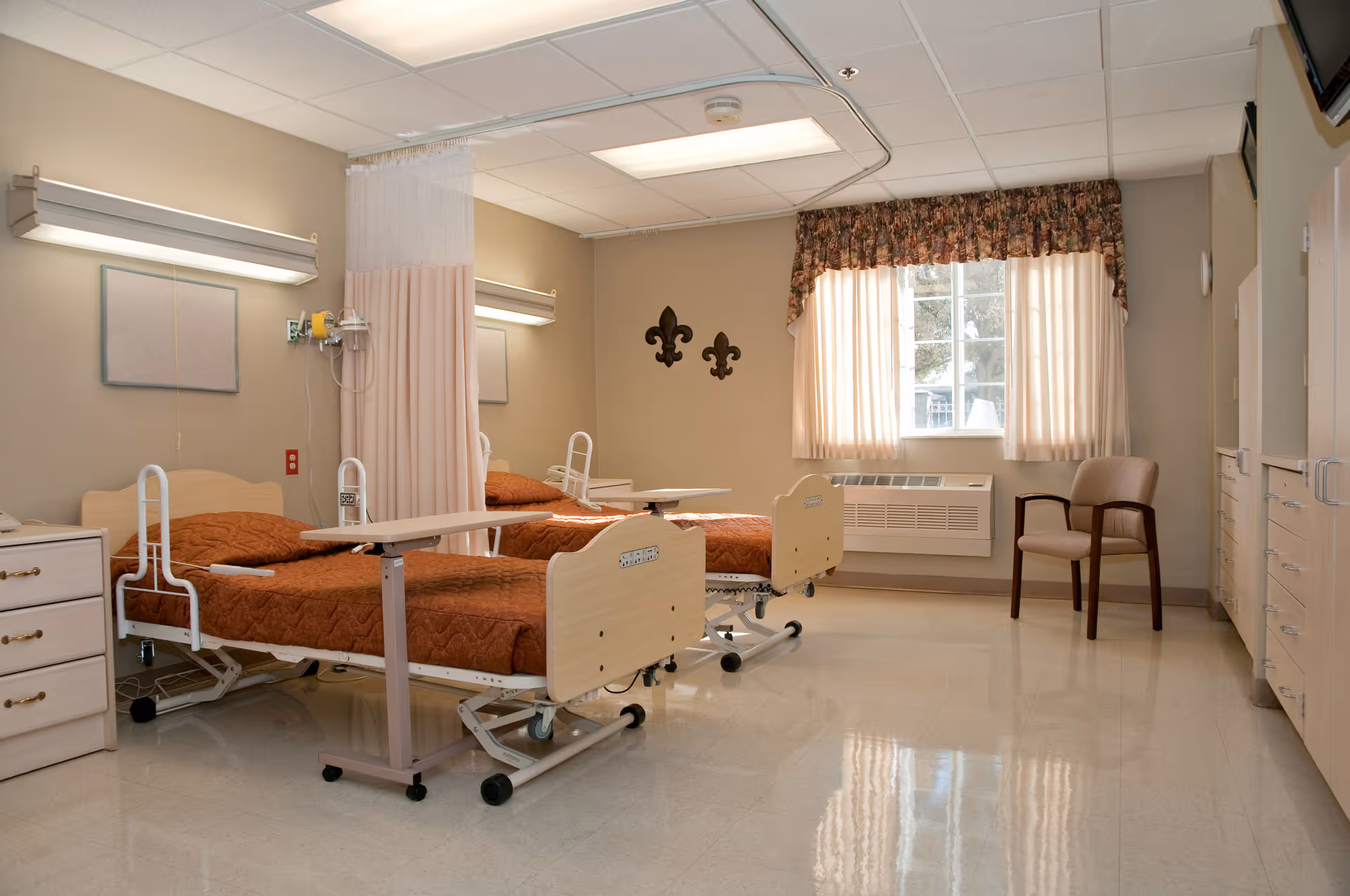 A clean and well-lit room with two hospital-style beds separated by a privacy curtain. Each bed has a brown quilted blanket and an adjustable overbed table. The room has beige walls, a window with floral valance and sheer curtains, a beige armchair, and medical equipment mounted on the wall. The floor is shiny and reflective.