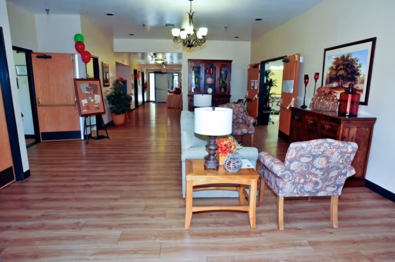 A spacious hallway in an assisted living facility with wooden flooring, a seating area including a patterned armchair, a small wooden table with a lamp and decorative items, and a sofa. The hallway has doors on both sides, potted plants, framed artwork on the walls, and ceiling light fixtures.