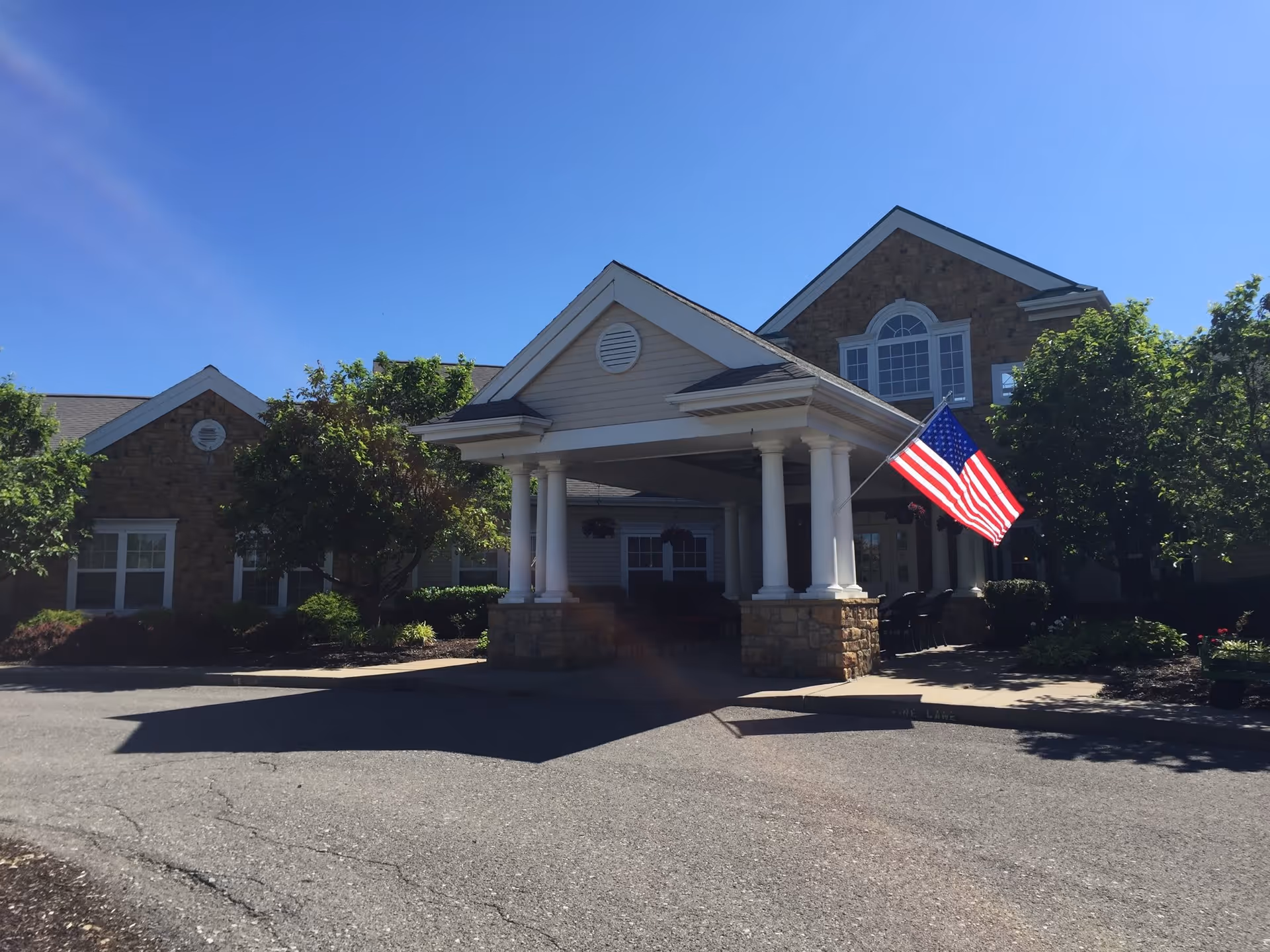 Front exterior view of a senior living facility building with a covered entrance supported by white columns. An American flag is displayed near the entrance. The building has stone and siding exterior with several windows and is surrounded by trees and landscaping under a clear blue sky.
