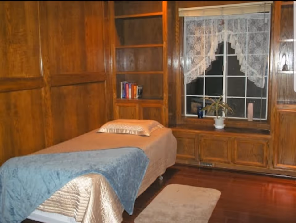 A cozy room with wooden paneled walls and built-in shelves. There is a single bed covered with a beige bedspread and a blue blanket at the foot. A pillow rests at the head of the bed. A window with lace curtains is above a wooden bench, which holds a potted plant and two small decorative items. The floor is wooden with a small beige rug beside the bed.