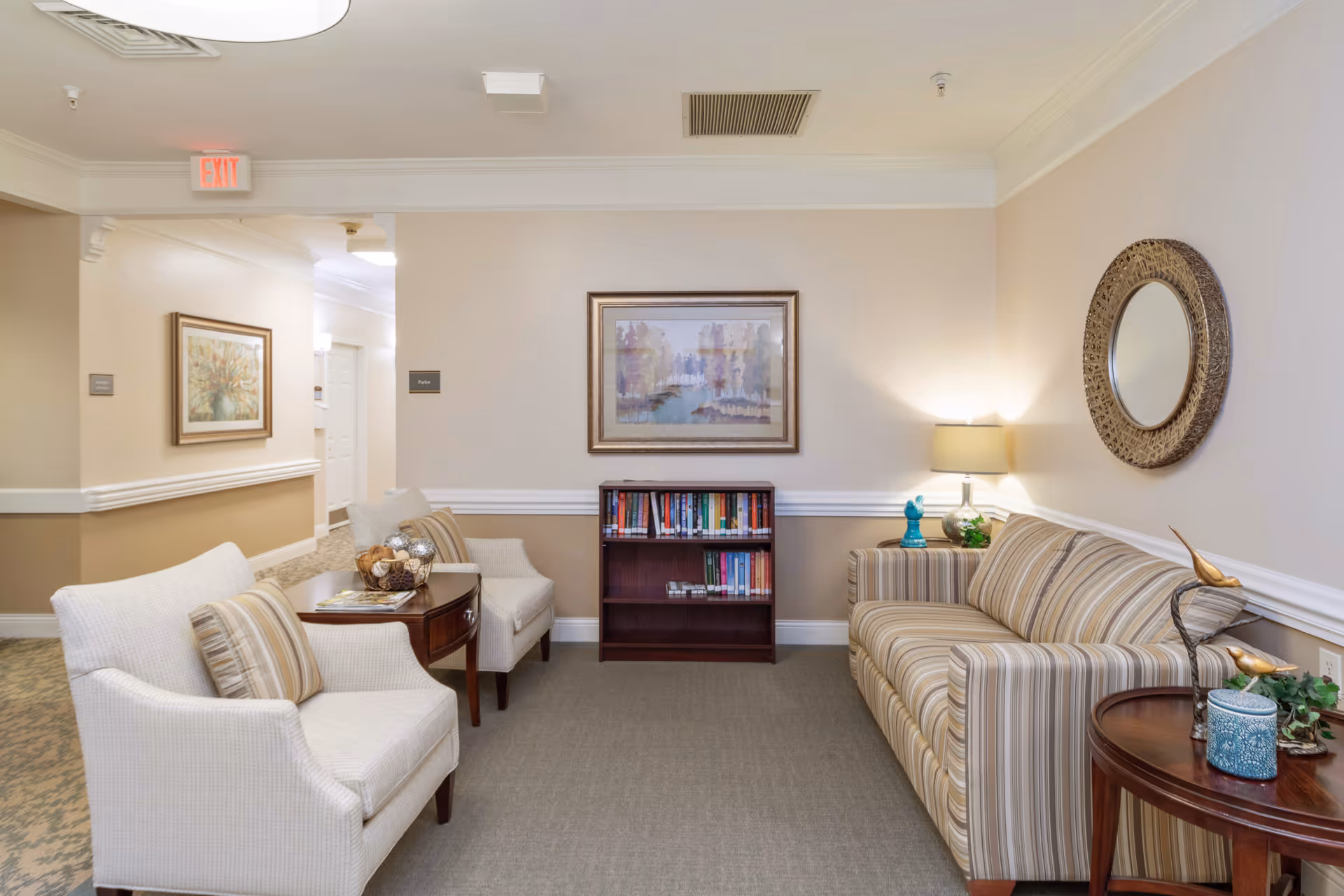 A comfortable senior living lounge with two armchairs, a striped sofa, a small bookshelf, and wall art.