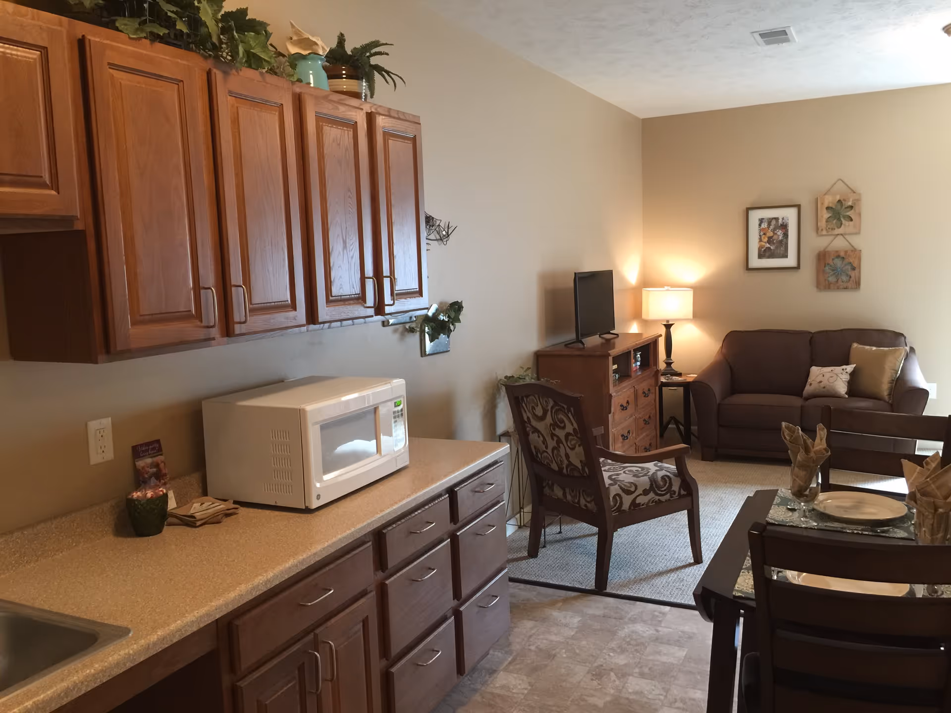 Interior view of a senior living facility room showing a kitchen area with wooden cabinets, a microwave on the countertop, and a small living area with a brown couch, a patterned armchair, a wooden TV stand with a flat screen TV, a table lamp, and wall decorations. A dining table with place settings is partially visible in the foreground.