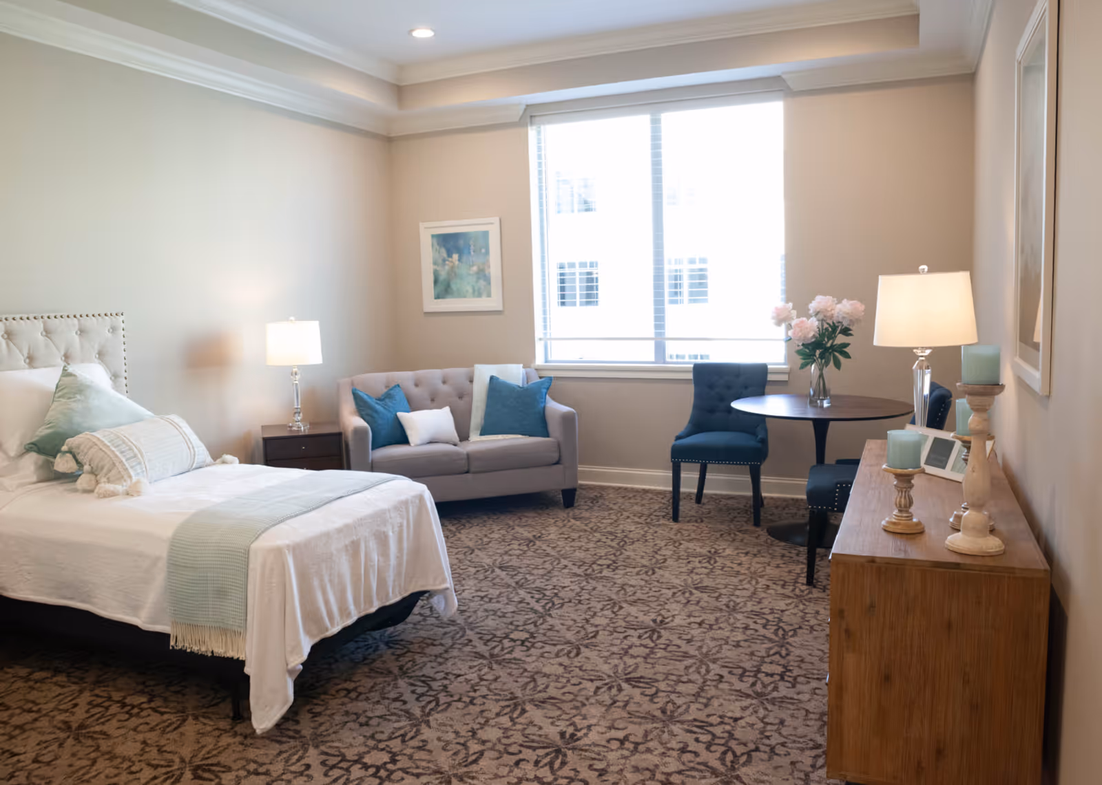 A well-lit bedroom in a senior living facility featuring a bed with white linens and light green pillows, a small gray loveseat with blue and white cushions, a round wooden table with two blue upholstered chairs, a wooden dresser with decorative candles and a lamp, and a large window with blinds letting in natural light.