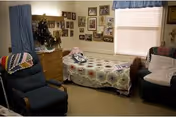 A cozy bedroom in a nursing home with a single bed covered by a patterned quilt, two armchairs with cushions, a wooden dresser, and a wall decorated with numerous framed photos. A window with blinds and a blue valance lets in natural light.