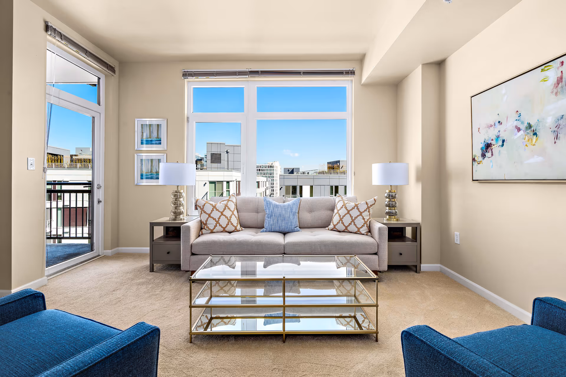 A bright living room with a beige sofa adorned with three decorative pillows, two side tables each with a lamp, a glass coffee table with gold trim, two blue armchairs, large windows showing a cityscape, and a colorful abstract painting on the wall.