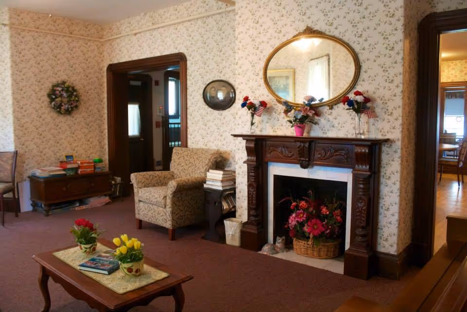 A cozy living room with floral wallpaper, a wooden fireplace mantel decorated with flower vases and a large oval mirror above it. There is an upholstered armchair next to the fireplace with a stack of books on a small side table. A wooden coffee table with flower pots and books is in the foreground. The room has carpeted flooring and wooden door frames leading to other rooms.