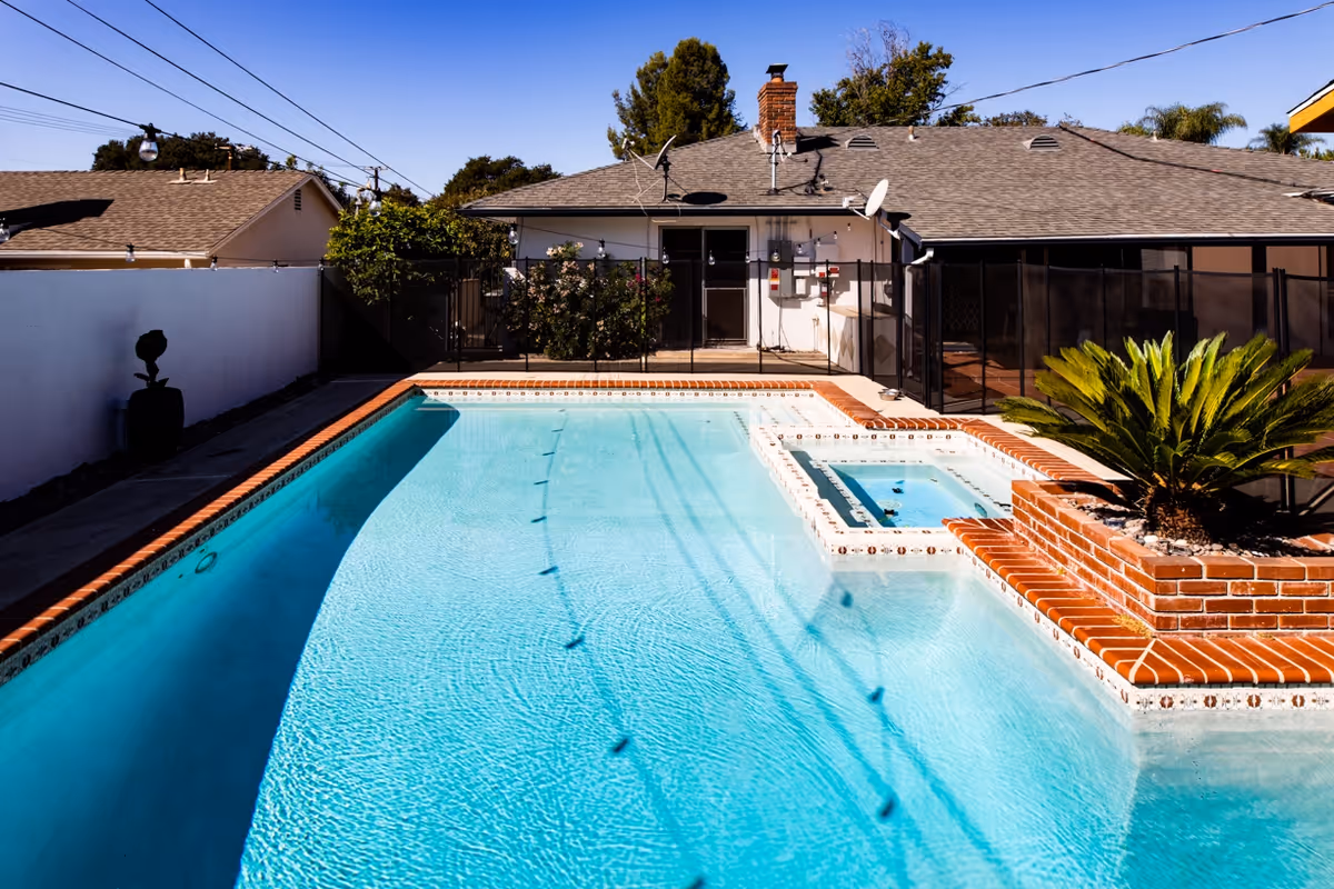 Outdoor swimming pool with clear blue water, adjacent hot tub, and a brick planter with a green plant. The pool area is surrounded by a black safety fence, and a single-story house with a chimney and satellite dishes is visible in the background under a clear blue sky.