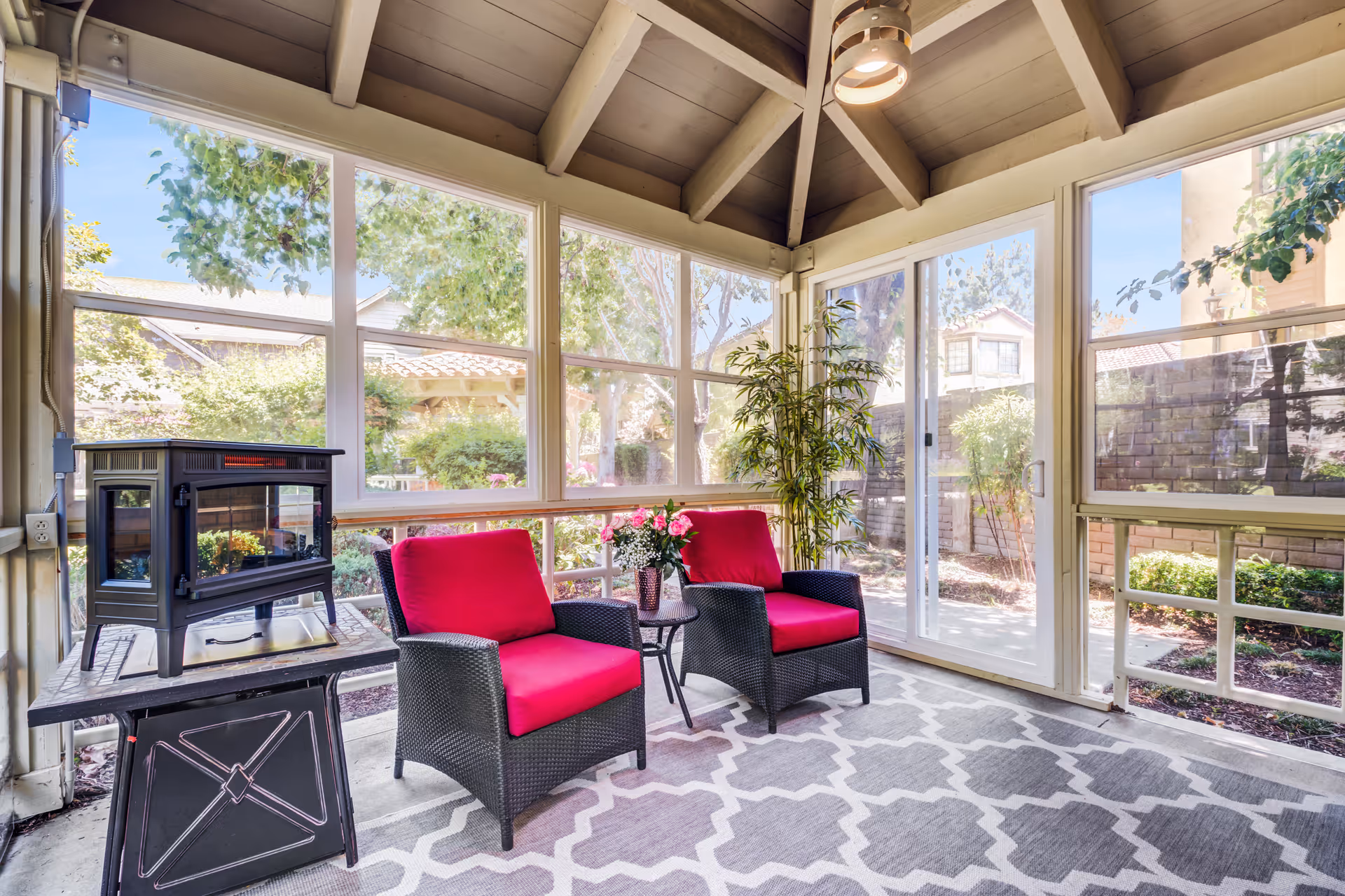 A bright enclosed patio area with large windows and sliding glass door showing an outdoor garden. Inside, there are two black wicker chairs with bright red cushions, a small round table with a flower vase, a patterned gray and white rug, a potted plant, and a black electric fireplace on a stand. The ceiling is vaulted with exposed beams and a hanging light fixture.