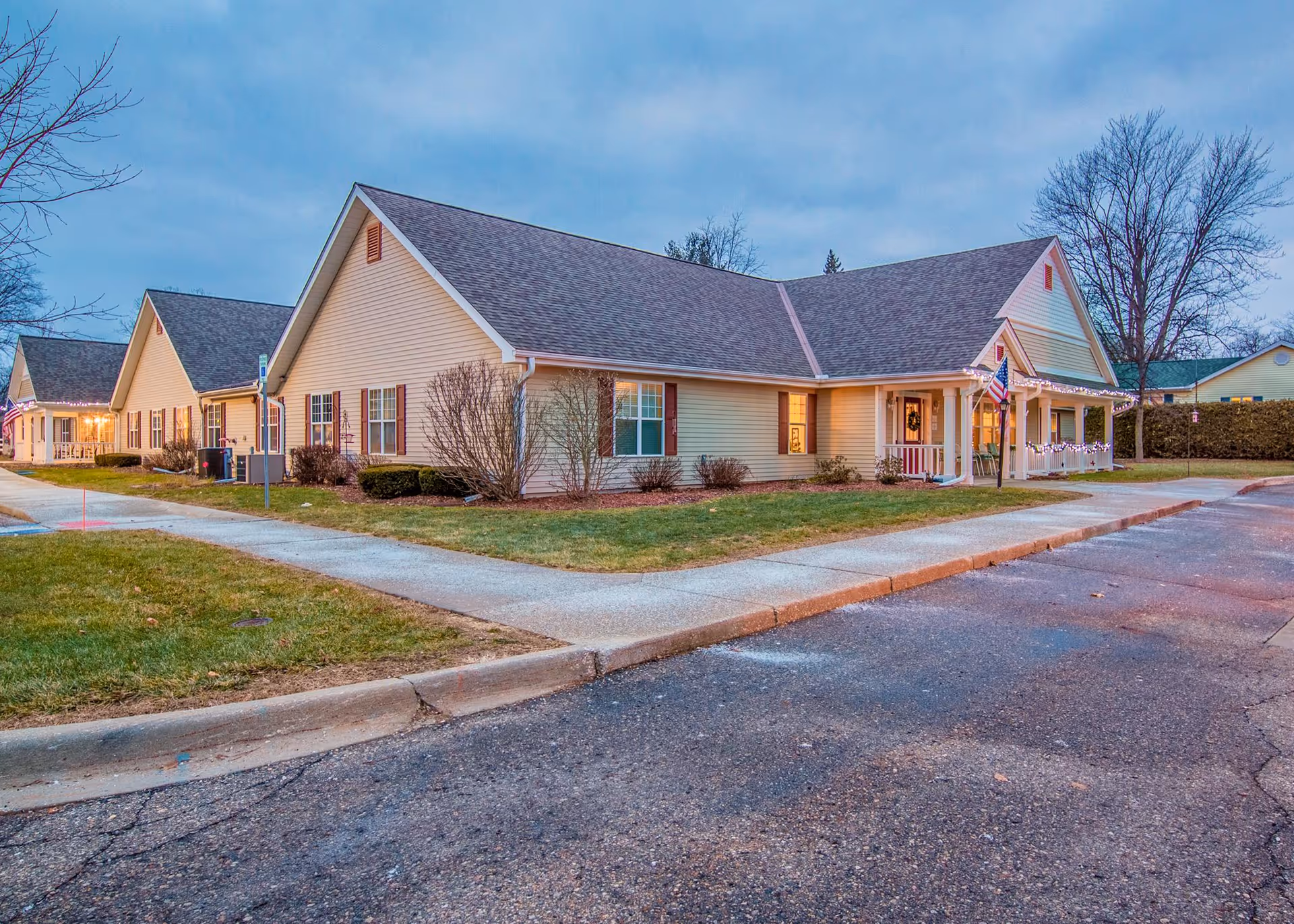 Exterior view of a single-story senior living facility building with beige siding, a gray shingled roof, and a small porch decorated with lights and an American flag. The building is surrounded by a sidewalk, grass, and some leafless trees under a cloudy sky at dusk.