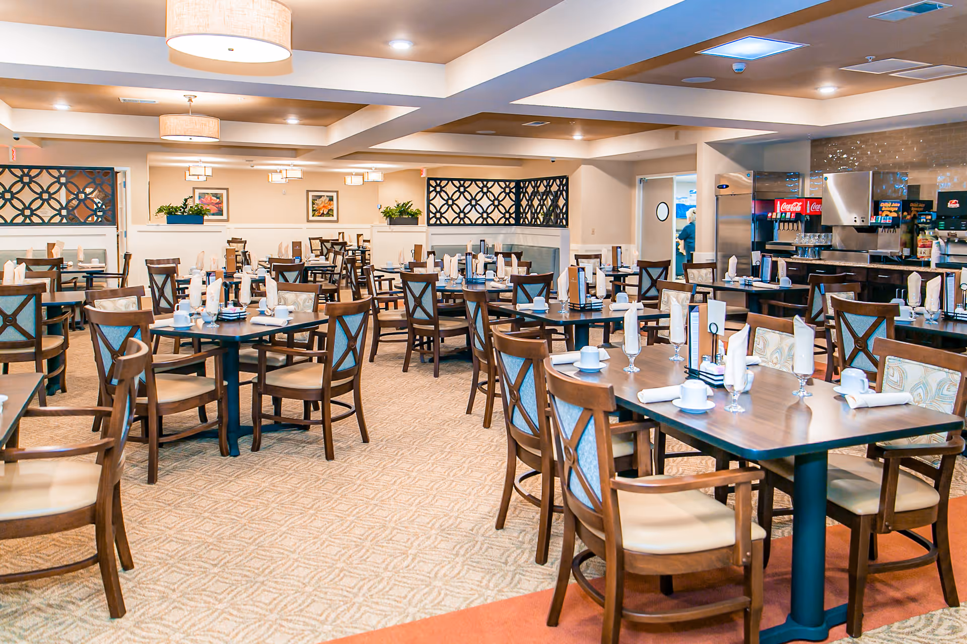 A spacious dining room in a senior living facility with multiple wooden tables and chairs arranged neatly. Each table is set with white napkins, cups, glasses, and menus. The room has warm lighting with ceiling lamps and decorative wall panels. In the background, there is a beverage station with soda dispensers and a staff member visible through a door.