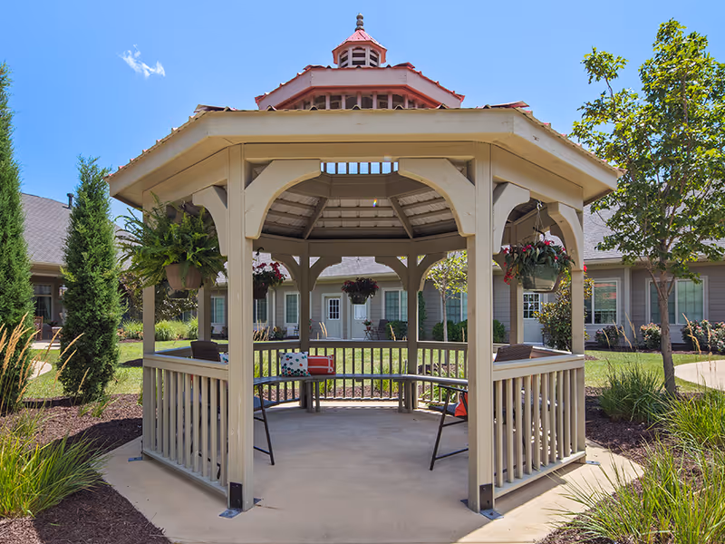 A beige wooden gazebo with a red roof situated in a landscaped garden area with green grass, trees, and shrubs. The gazebo has hanging flower pots and benches inside, with a building featuring multiple windows and doors visible in the background under a clear blue sky.
