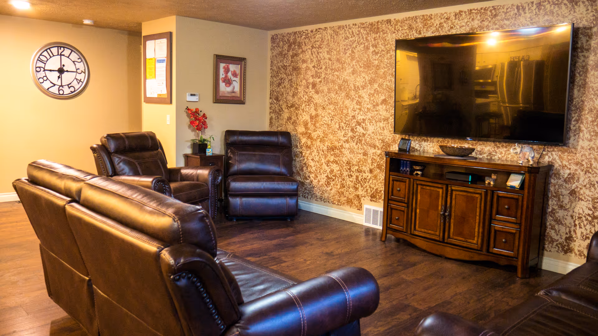 A cozy living room with brown leather sofas and armchairs arranged around a large flat-screen TV mounted on a textured wall. A wooden TV stand holds decorative items and electronics. The room has wooden flooring, a wall clock, framed artwork, and a small table with a red flower arrangement.