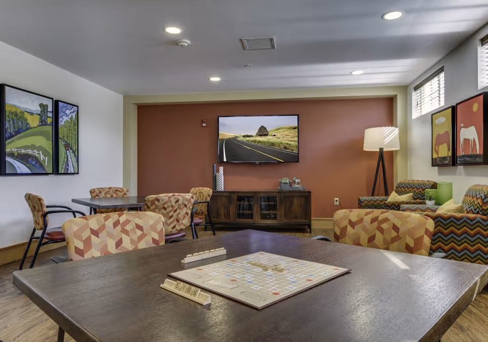 A comfortable senior lounge with tables and patterned chairs, a TV on a red accent wall, and a Scrabble game on the foreground table.