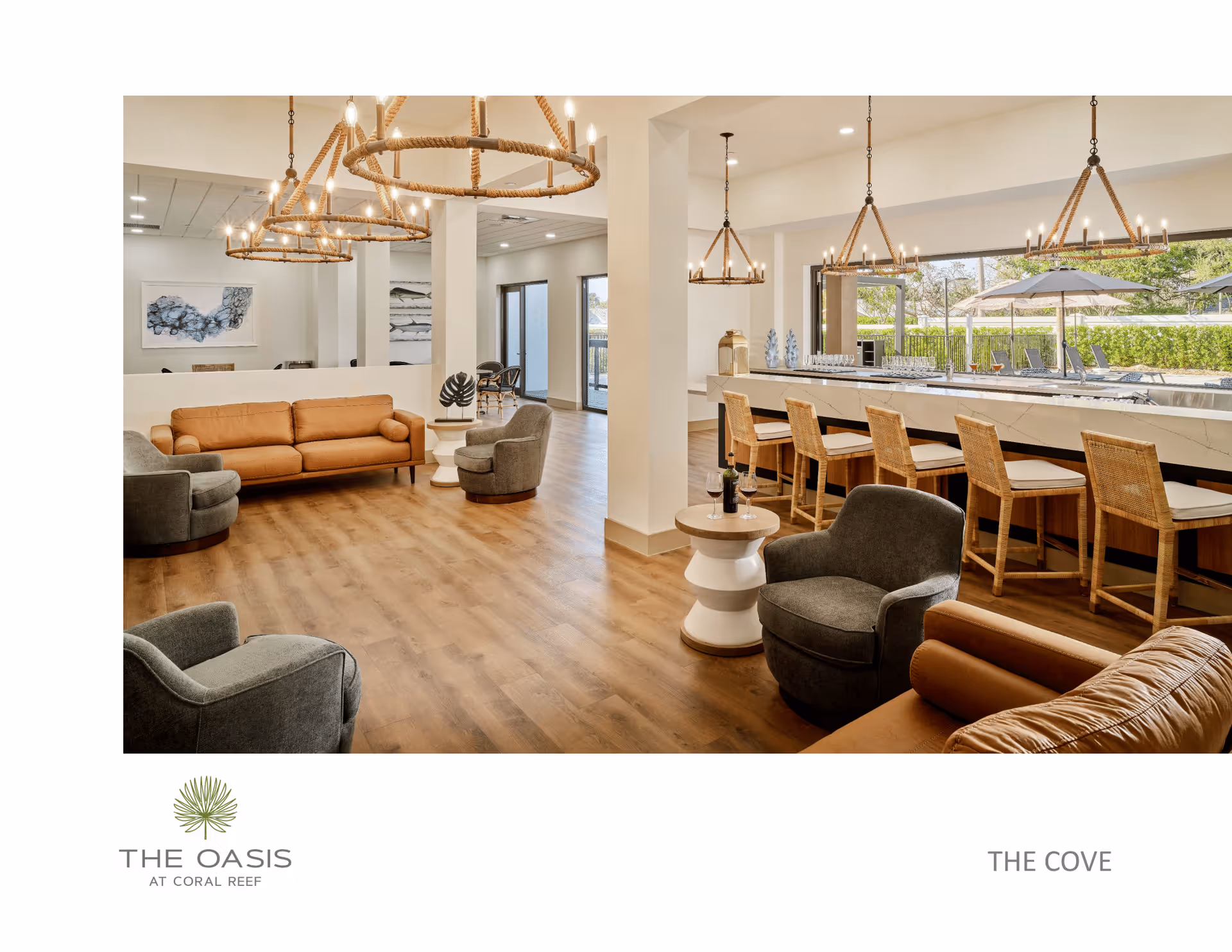 A spacious and modern common area with wooden flooring, featuring a seating area with a tan sofa and gray armchairs, a bar counter with wicker bar stools, and large windows showing an outdoor patio with umbrellas and lounge chairs. The room is illuminated by multiple rustic rope chandeliers.