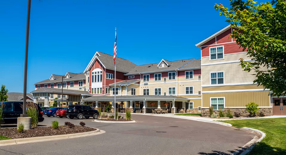 Exterior view of a large senior living facility building with red, beige, and yellow siding under a clear blue sky. There is a driveway with parked cars, a flagpole with an American flag, and some landscaping with trees and shrubs.
