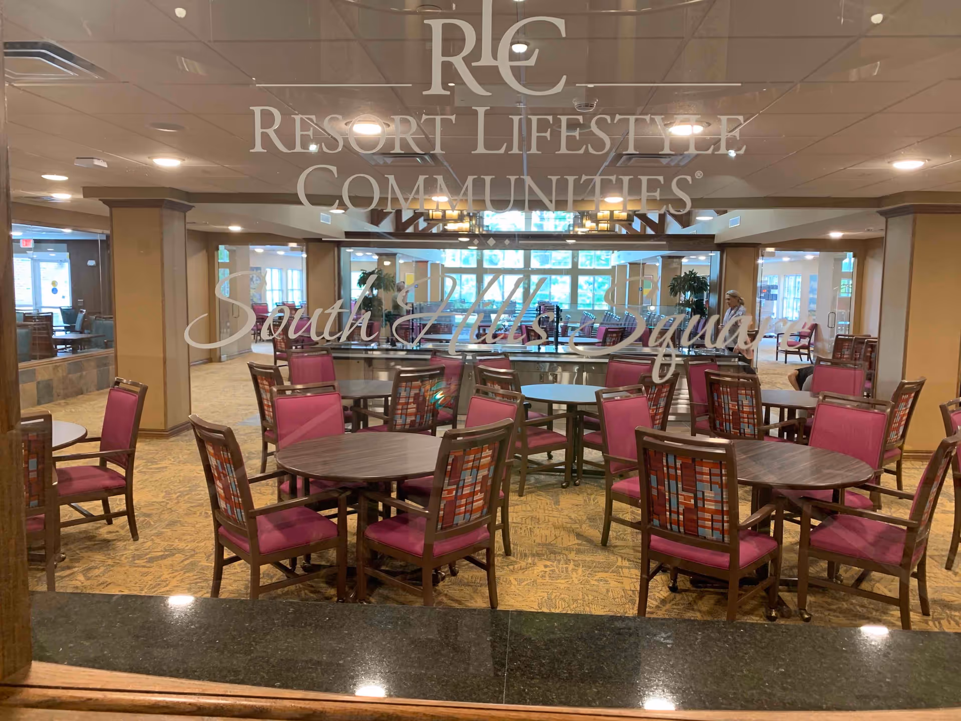 Interior view of a dining area with multiple round tables and chairs with pink cushions and patterned backs. The room has a carpeted floor, large windows letting in natural light, and a few plants. The image is taken through a glass panel with the text 'Resort Lifestyle Communities South Hills Square' etched on it.