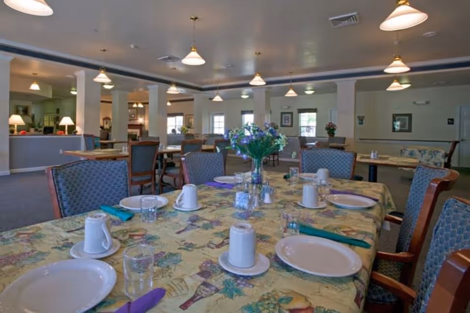 A dining room in a senior living facility with tables set for a meal. The tables have floral-patterned tablecloths, plates, cups, glasses, and napkins. There are upholstered chairs around the tables and a vase with flowers as a centerpiece. The room is well-lit with hanging pendant lights and has multiple windows and columns.