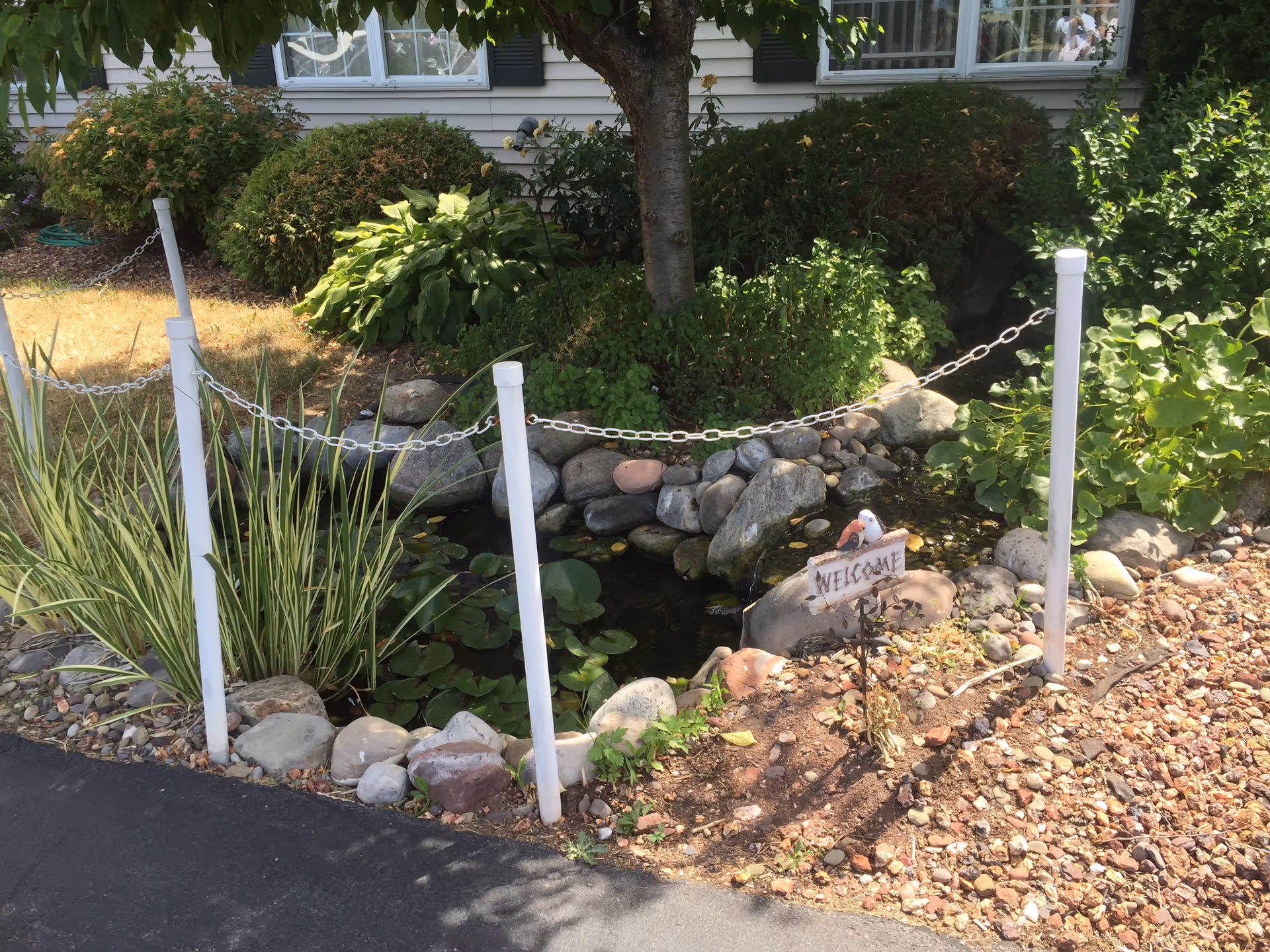 Small landscaped front-yard pond with rocks, plants, a 'Welcome' sign and white chain posts in front of a building.