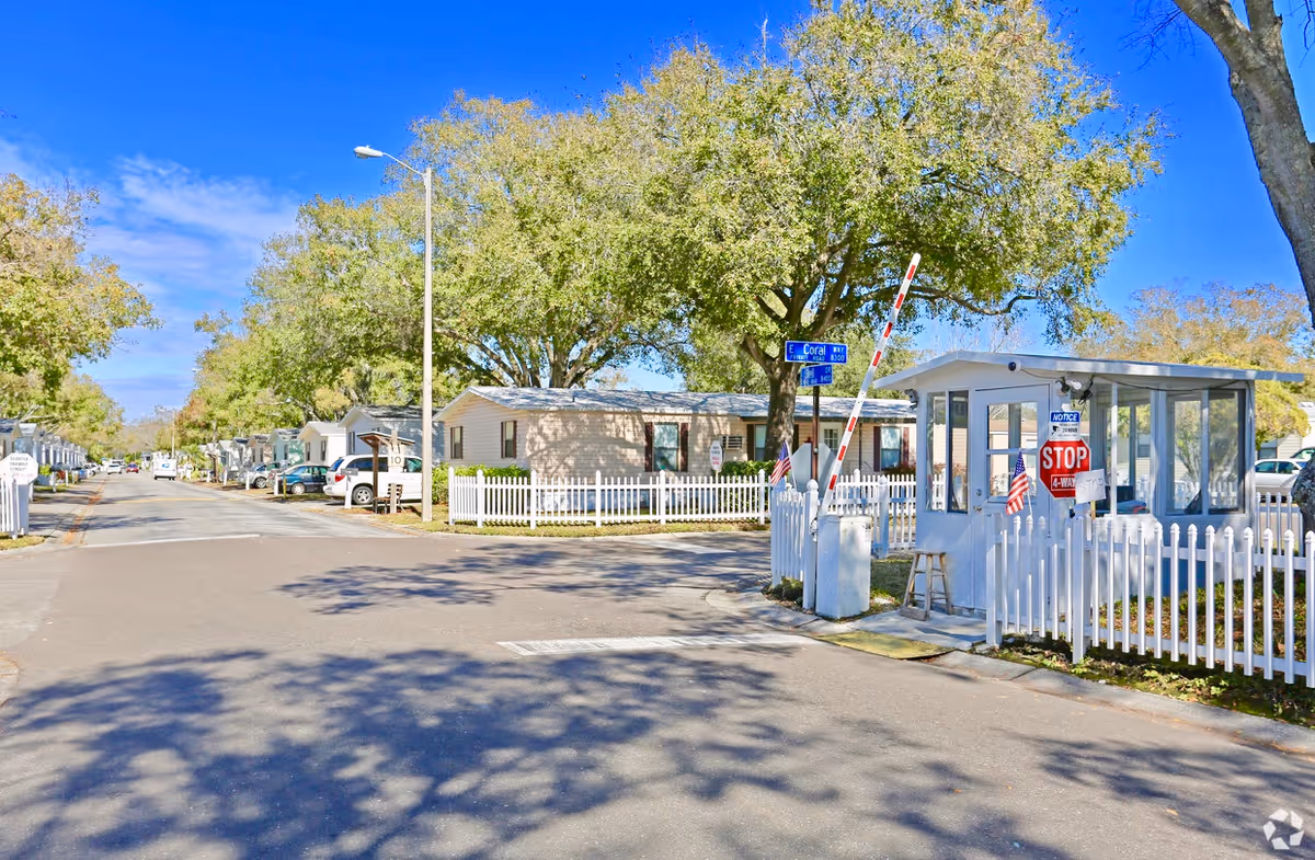 Entrance to a gated, mobile-home style community with a guard booth, white picket fences and a tree-lined street.