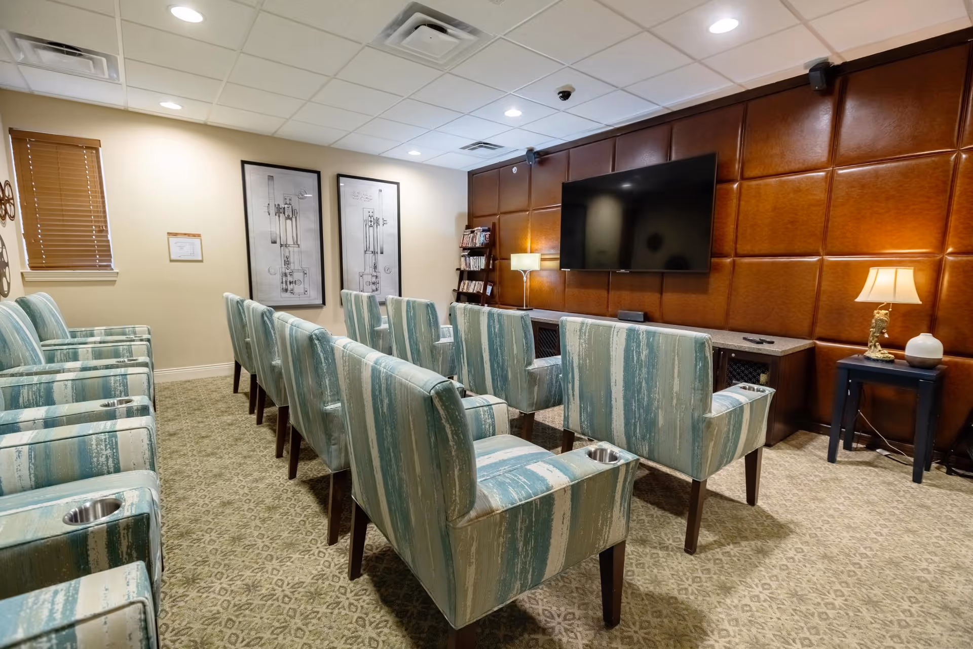 Small media room with rows of blue-green upholstered chairs facing a wall-mounted TV on a leather-paneled accent wall.