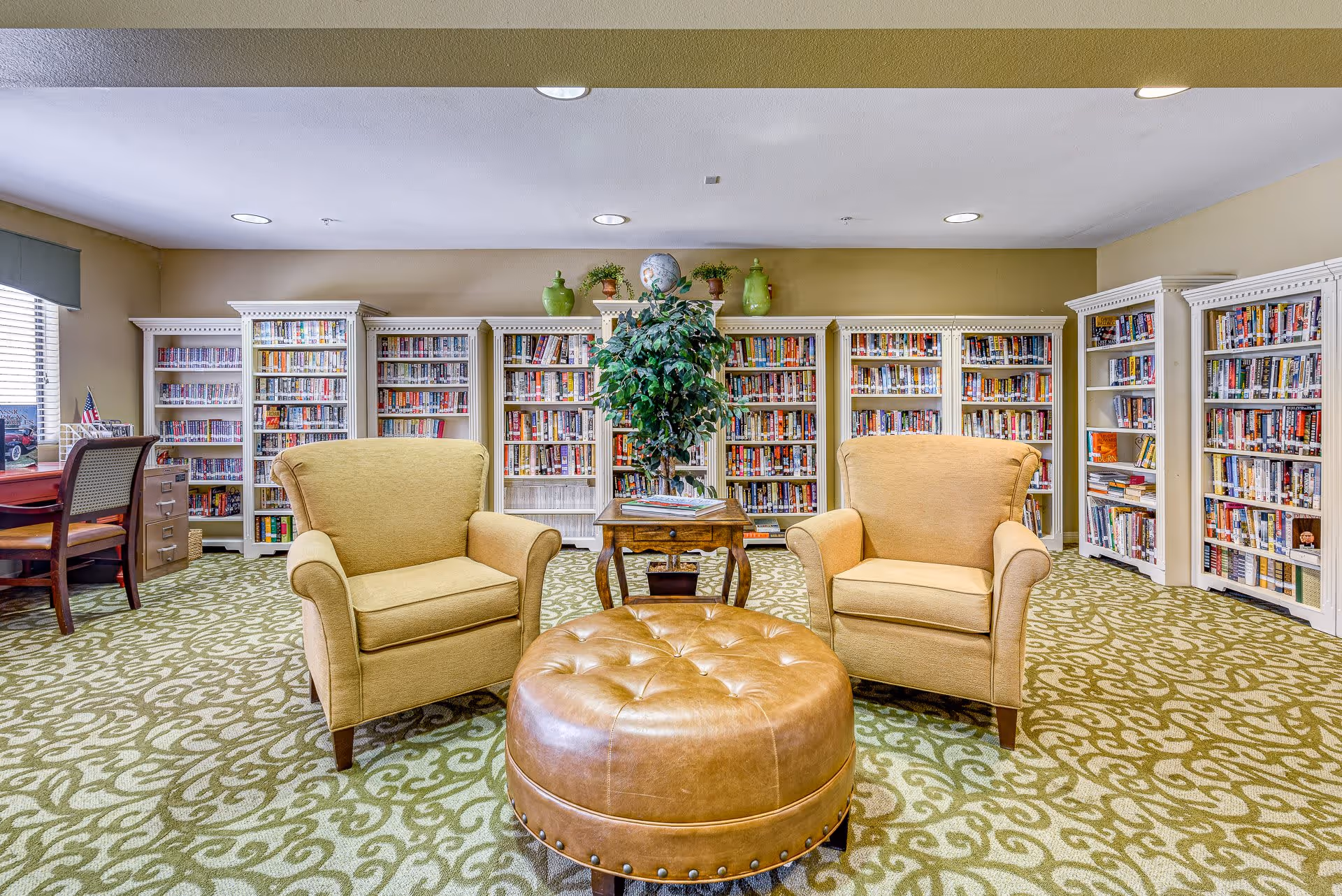 A cozy library room with two beige armchairs and a round brown leather ottoman in the center. Behind the chairs are white bookshelves filled with books, and a small wooden table with a potted plant on top. The room has patterned green carpet and beige walls with recessed lighting in the ceiling.