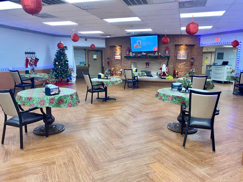 A festive dining area decorated for Christmas with round tables covered in green and red holiday-themed tablecloths, chairs around the tables, a decorated Christmas tree, hanging red ornaments from the ceiling, and a wall-mounted TV above a fireplace mantel adorned with holiday decorations including stockings and Santa figurines.