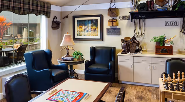 A cozy interior common area with two dark blue armchairs, a round side table with a lamp and plant, a large window showing an outdoor patio with chairs, a framed picture on the wall, and a cabinet with decorative items including a saddle and a chessboard on a table in the foreground.