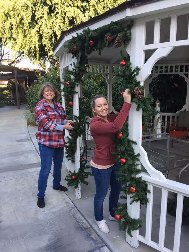 Two women decorating a white gazebo outdoors with green garlands adorned with red ornaments and pinecones. One woman is wearing a red plaid shirt and jeans, and the other is wearing a maroon hoodie and jeans. The setting appears to be a garden or courtyard area with trees and shrubs in the background.