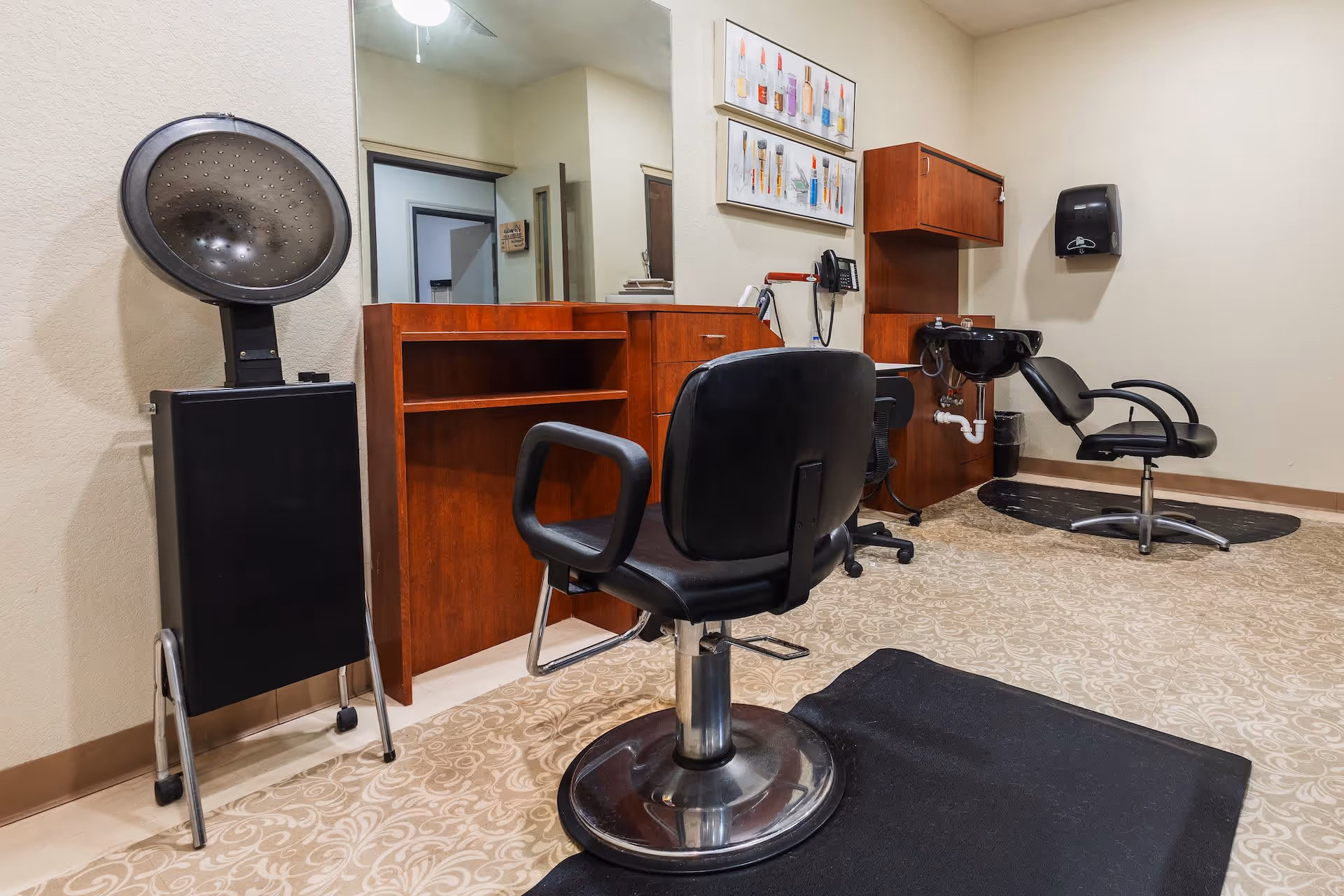 Interior view of a senior living facility hair salon with two black salon chairs, a hair dryer, a wooden cabinet with a large mirror, a black sink for washing hair, and a wall-mounted paper towel dispenser. The floor has a patterned carpet and there are framed pictures on the wall.
