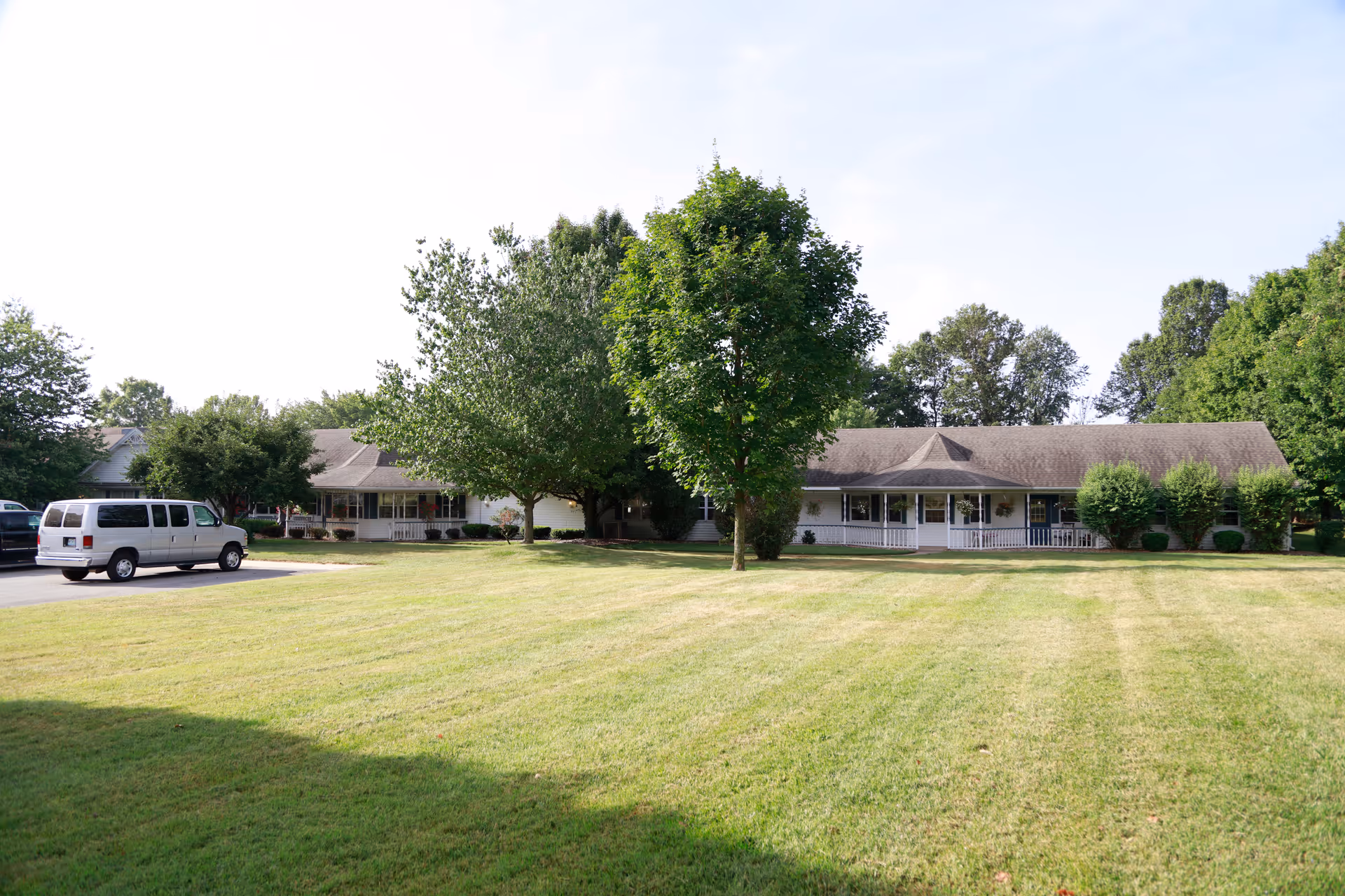 A single-story building with a large front porch surrounded by trees and a well-maintained grassy lawn. A white van is parked on the driveway to the left of the building. The sky is clear with some light clouds.