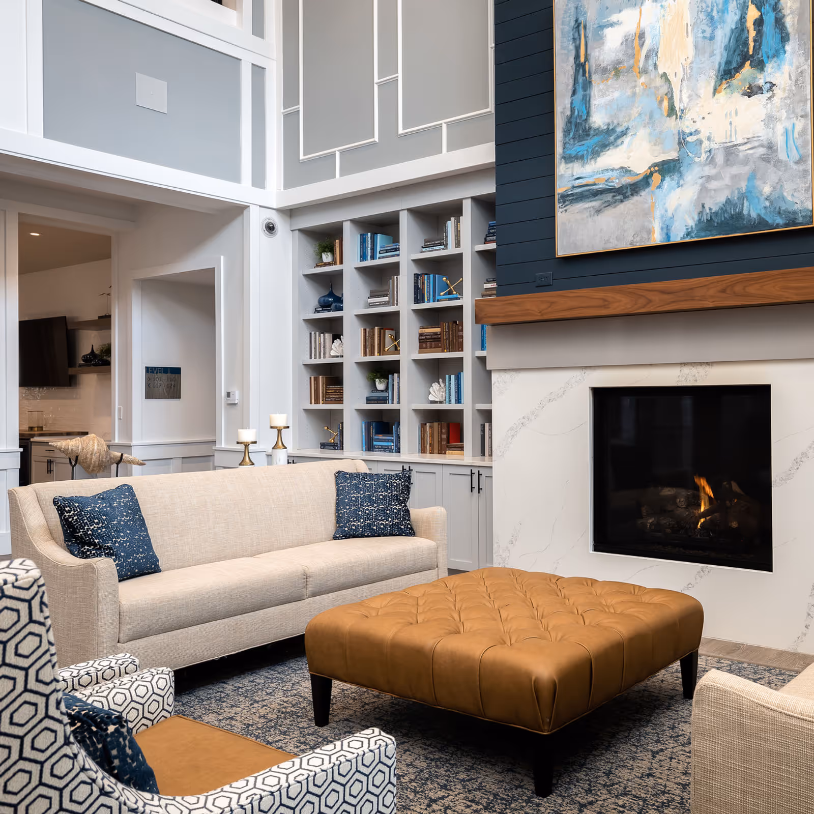 A stylish living room with beige sofas, a tufted leather ottoman, built-in bookshelves, and a fireplace beneath abstract artwork.