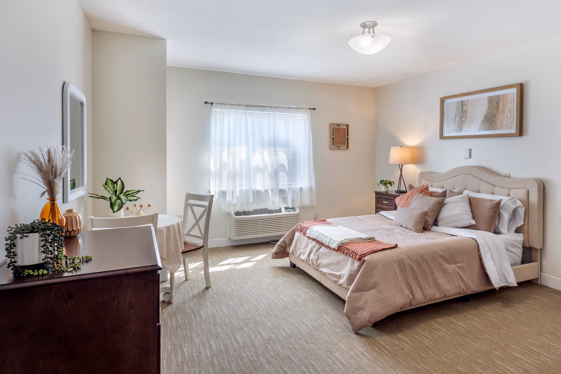 A bright and cozy bedroom with a large bed adorned with multiple pillows and a beige comforter. There is a nightstand with a lamp and a small plant beside the bed. A window with sheer white curtains allows natural light to fill the room. Across from the bed, there is a small round table with two chairs and a dresser with decorative plants and vases. The walls are light-colored, and a framed artwork hangs above the bed.