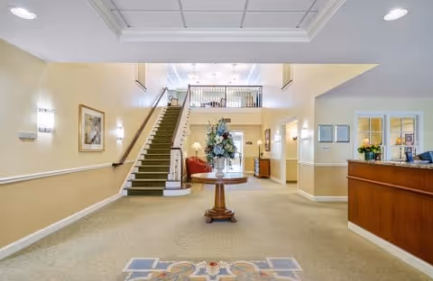 Spacious assisted living lobby with a central round table and floral arrangement, reception desk on the right, and a carpeted staircase leading to an upper level.