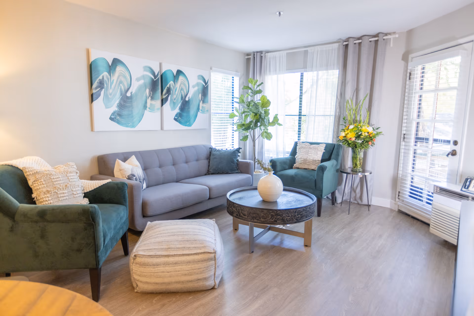 A bright and cozy living room with a gray sofa, two green armchairs, a round coffee table with a white vase, a pouf, and large windows with sheer curtains letting in natural light. There is also a side table with a vase of flowers and abstract artwork on the wall.
