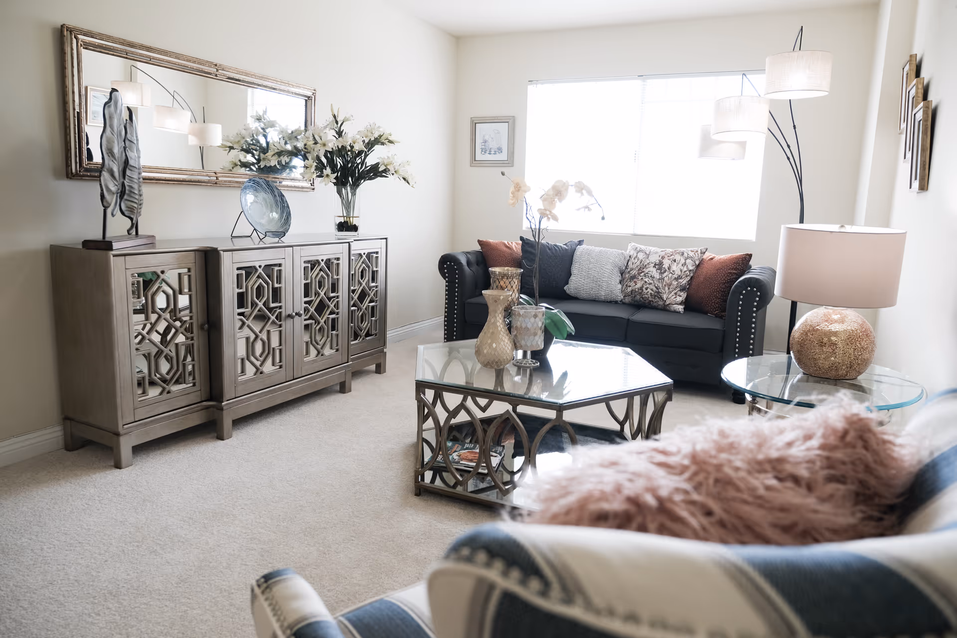 A bright living room with a dark sofa adorned with multiple cushions, a glass coffee table with decorative vases, a side table with a lamp, a large mirror above a wooden cabinet with decorative items, and a floor lamp near the window.