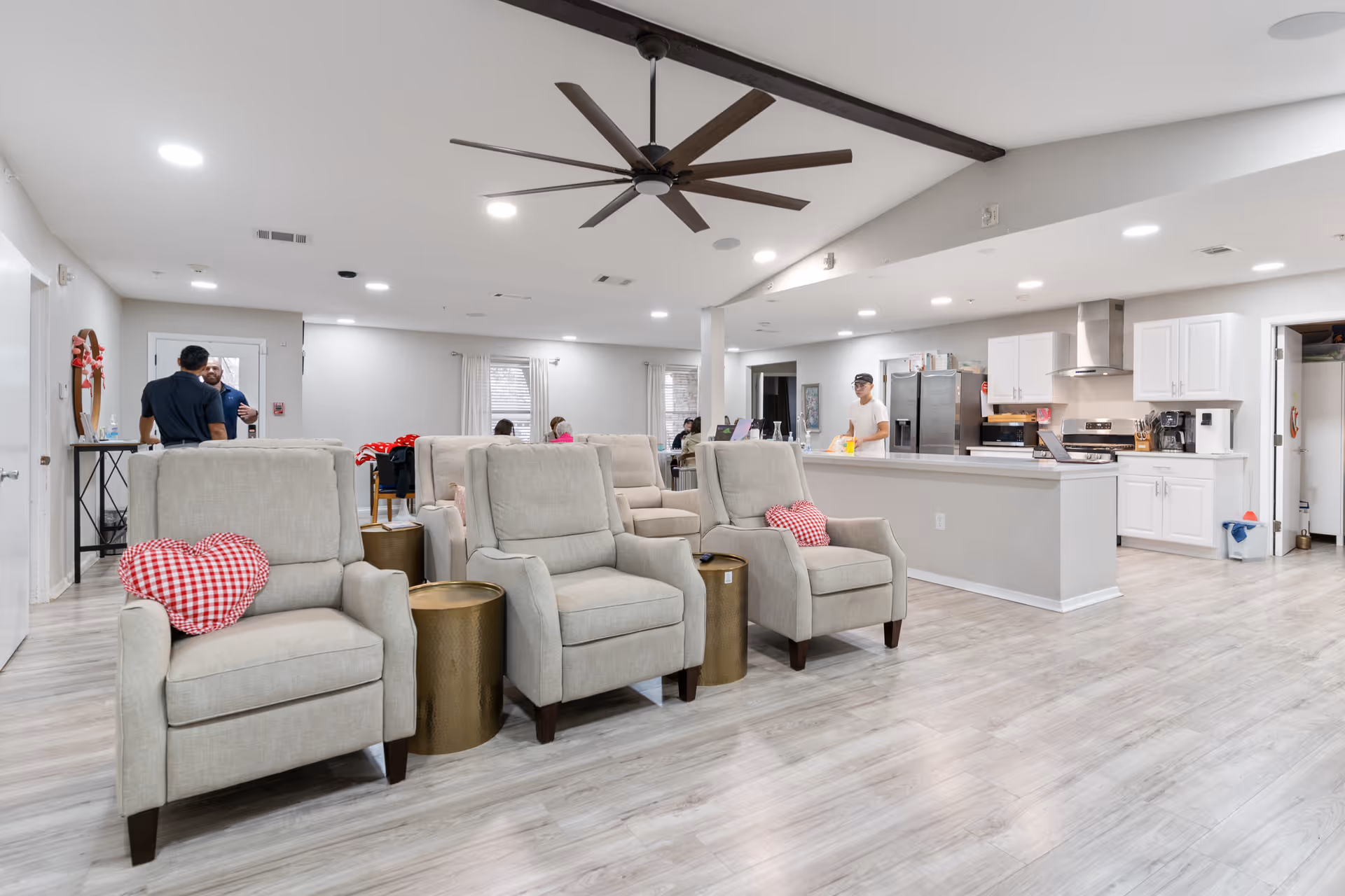 A spacious and bright common area in a senior living facility featuring three beige armchairs with red and white checkered heart-shaped pillows, two small round gold side tables, a large ceiling fan, and an open kitchen with white cabinets and stainless steel appliances. Several people are visible in the background near the kitchen and entrance area.