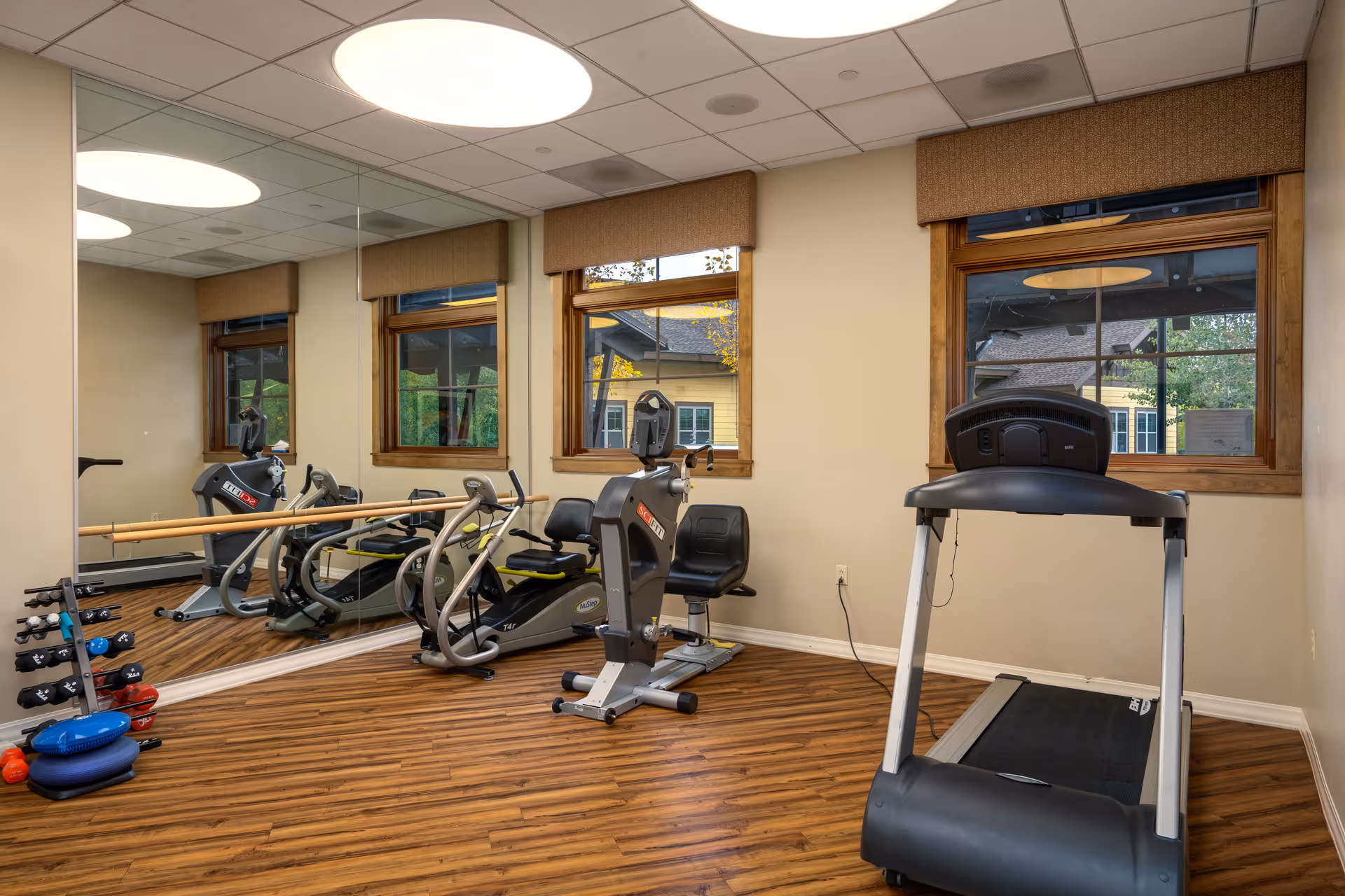 A small fitness room with wooden flooring, two windows with brown valances, a large wall mirror, a treadmill, a recumbent exercise bike, a rack of dumbbells, and some exercise equipment on the floor.