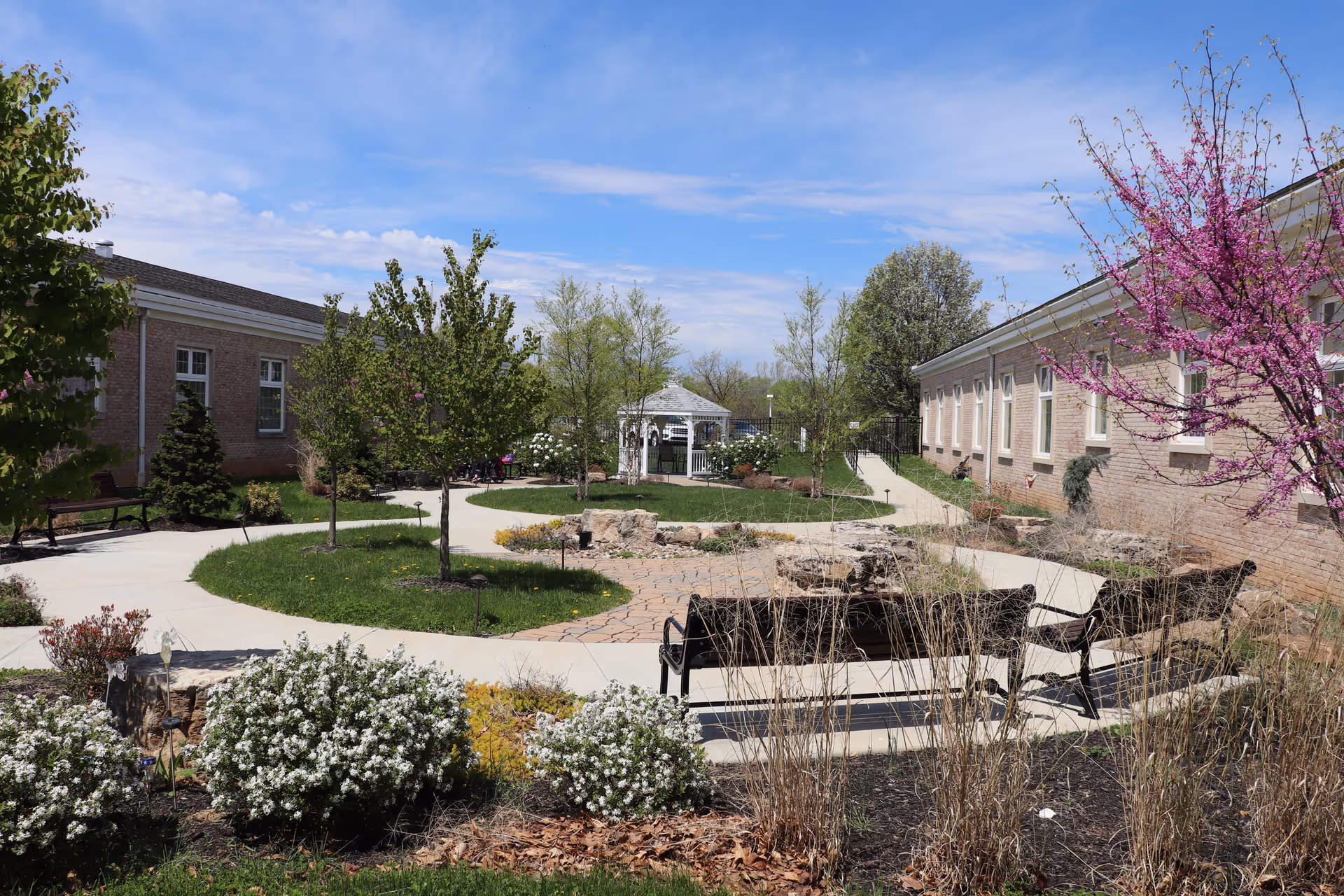 Outdoor garden area at Claremont Nursing & Rehabilitation Center featuring paved walkways, benches, flowering trees and shrubs, and a white gazebo in the background under a partly cloudy blue sky.
