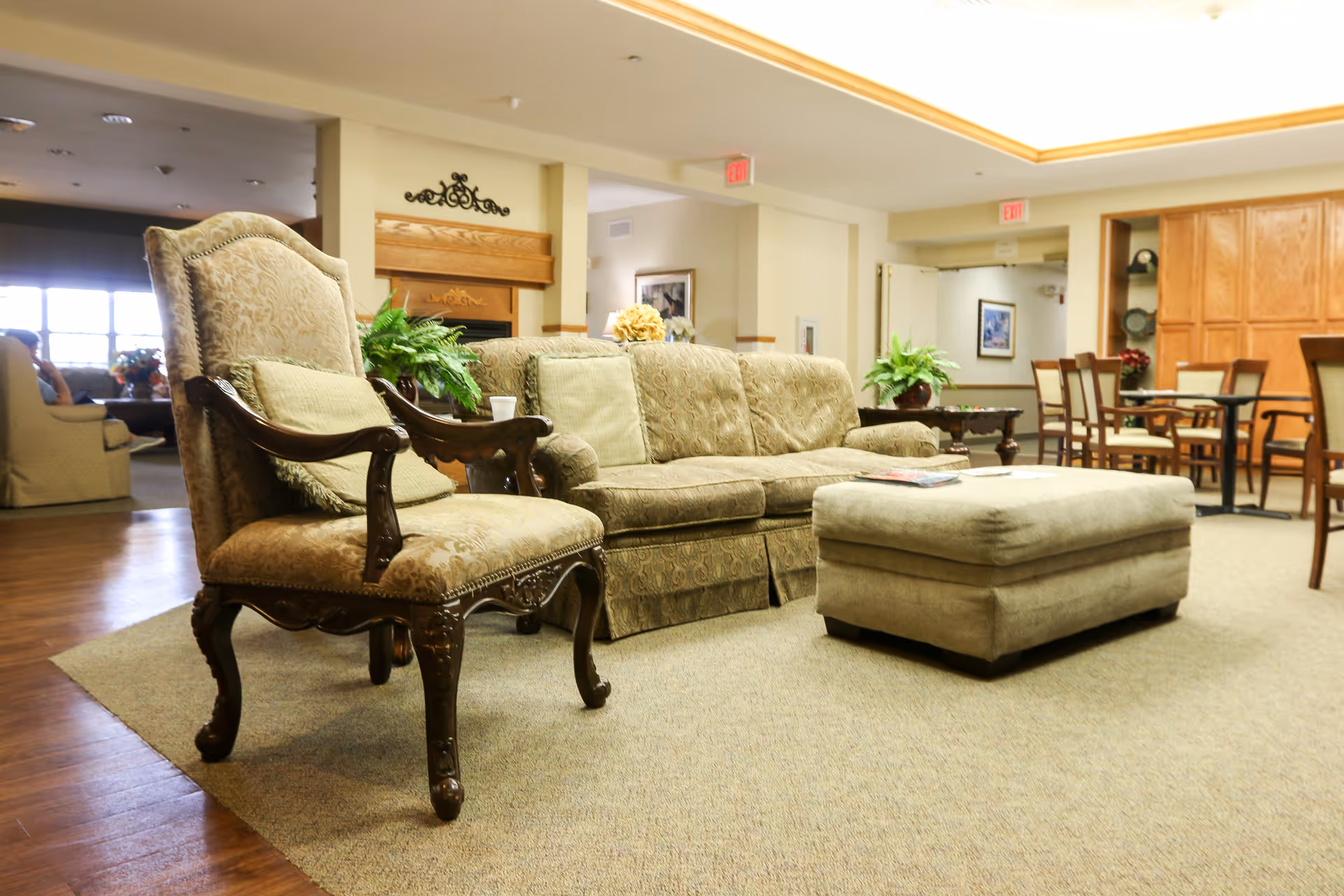 A cozy living room area in a senior living facility featuring a beige patterned armchair with wooden armrests, a matching sofa with cushions, and a large cushioned ottoman on a carpeted floor. In the background, there are wooden dining chairs and tables, potted plants, and warm lighting from ceiling fixtures.