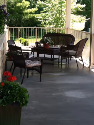 A covered outdoor patio area with wicker furniture including two chairs, a loveseat, and a table with a potted plant on it. The patio overlooks a green garden with trees and bushes in the background.
