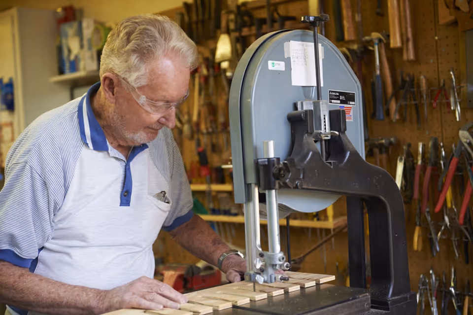 An elderly man wearing safety glasses operates a band saw to cut wooden pieces in a workshop filled with various hand tools hanging on a pegboard wall.