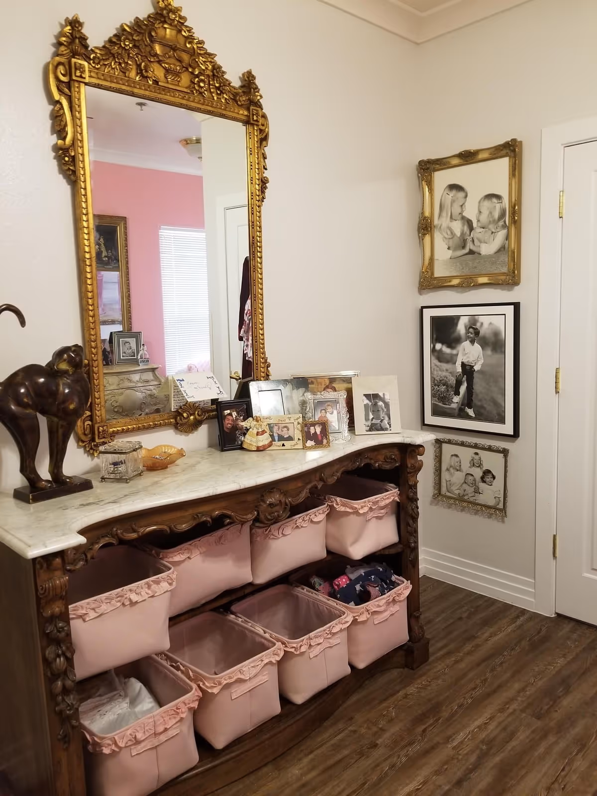 An ornate wooden console table with a white marble top holding several framed photos and decorative items. Above the table is a large, intricately carved gold-framed mirror reflecting part of a pink wall and window. Below the table are eight pink fabric storage bins. On the adjacent wall, there are three framed black and white photographs of children. The floor is wooden, and a white door is visible on the right side.