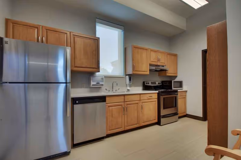 A modern kitchen with wooden cabinets, a stainless steel refrigerator, dishwasher, stove, and microwave. There is a window above the sink and the floor is tiled.