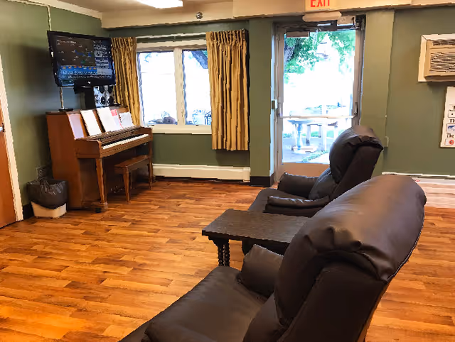 Communal sitting area with two leather recliners, a piano, a wall-mounted TV, and a glass exit door to the outside.